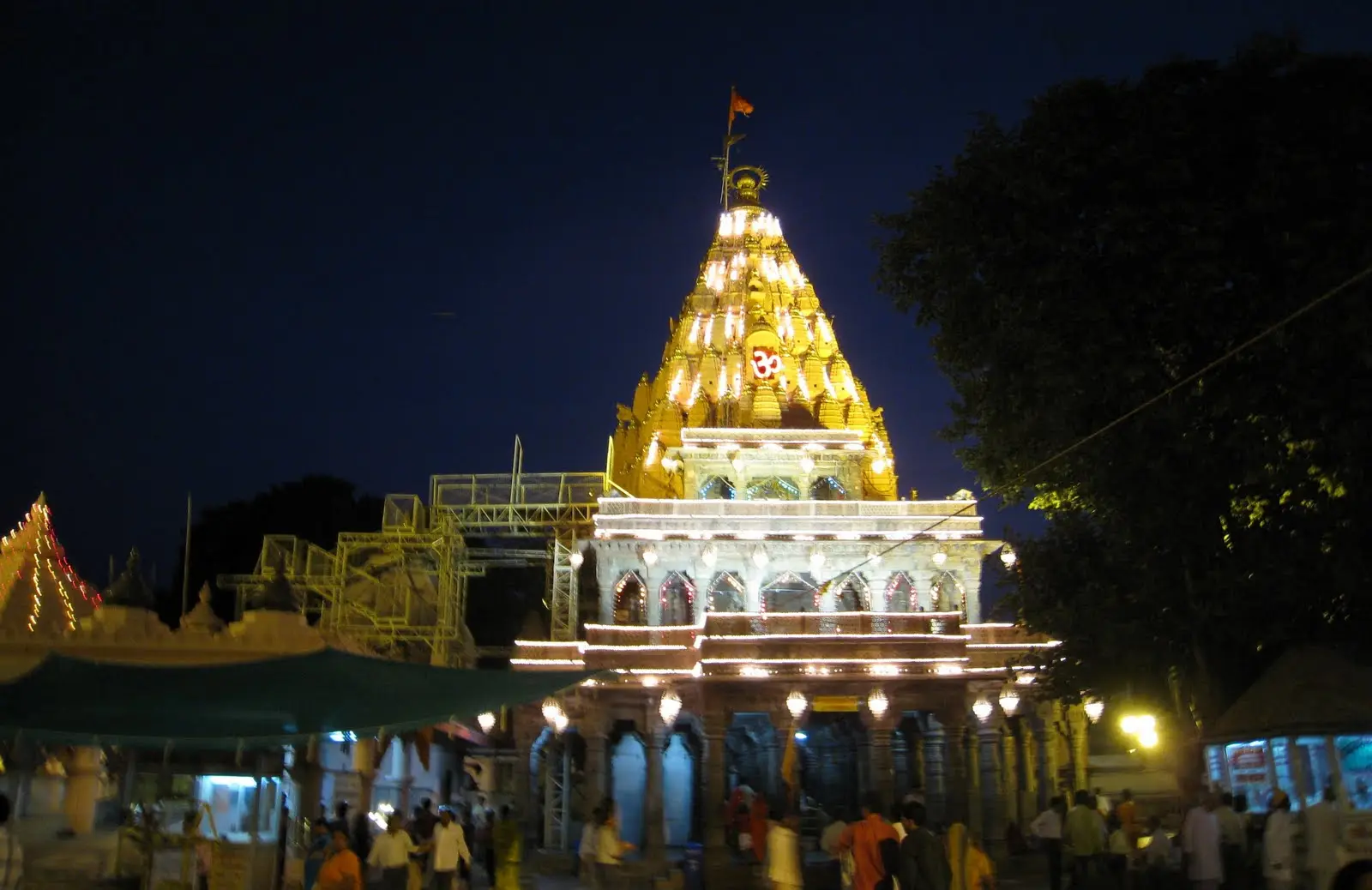 Mahakaleshwar temple in Ujjain at night, its shikhara lit gold with an Om sign, and devotees walking in the courtyard below