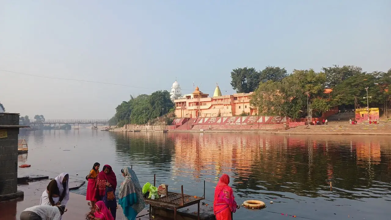 Devotees on the steps of Ram Ghat in Ujjain on a clear morning, with temples and trees on the far bank of the Shipra reflected in the water