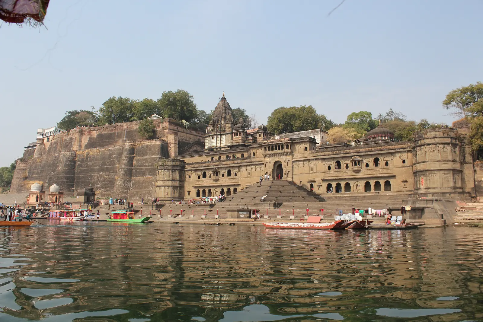Ahilya Fort's stone ramparts and palace front with the Ahilyeshwar temple shikhara at Maheshwar, seen across the Narmada with boats at the ghats