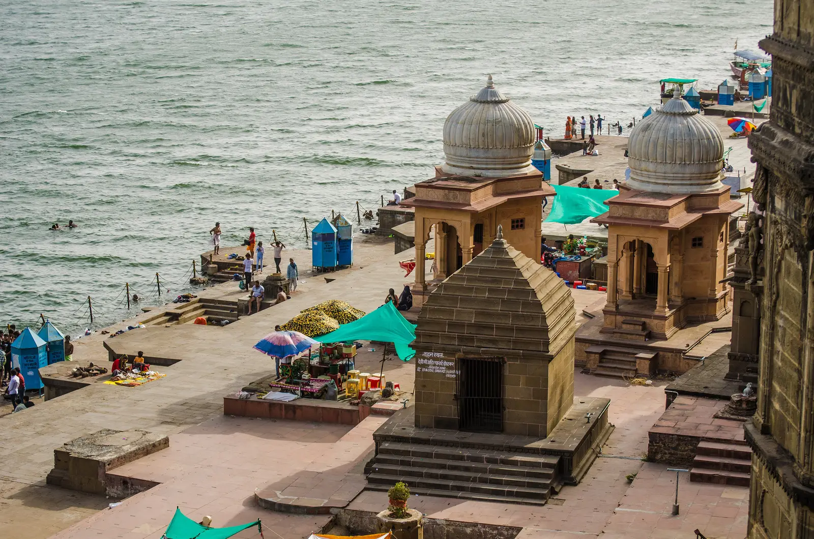 Overhead view of a Maheshwar ghat with two domed chhatris, a small stone shrine, stalls under umbrellas and bathers at the edge of the Narmada