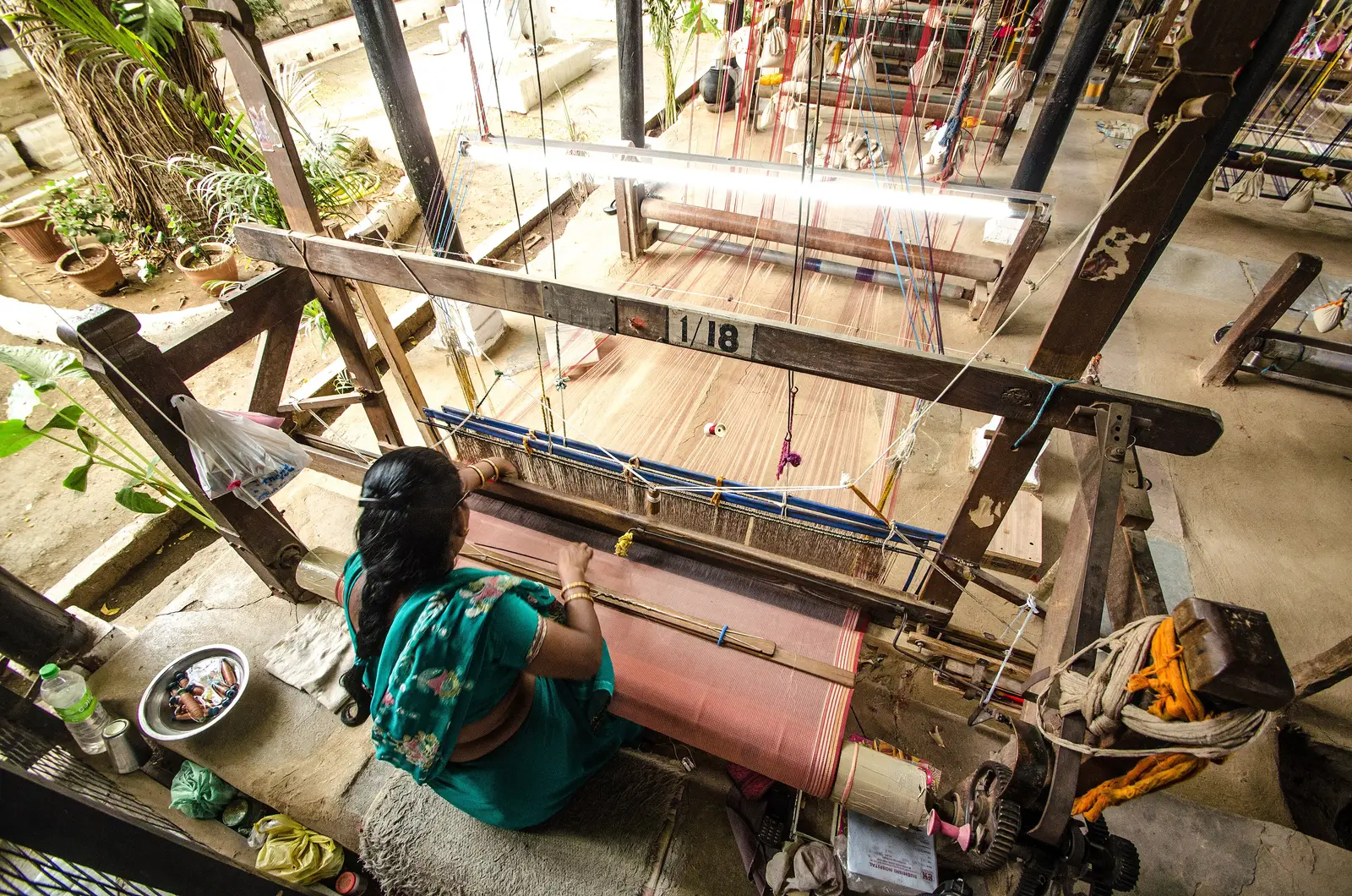 A weaver in a green sari working at a wooden handloom inside Maheshwar fort, with warp threads and other looms around her