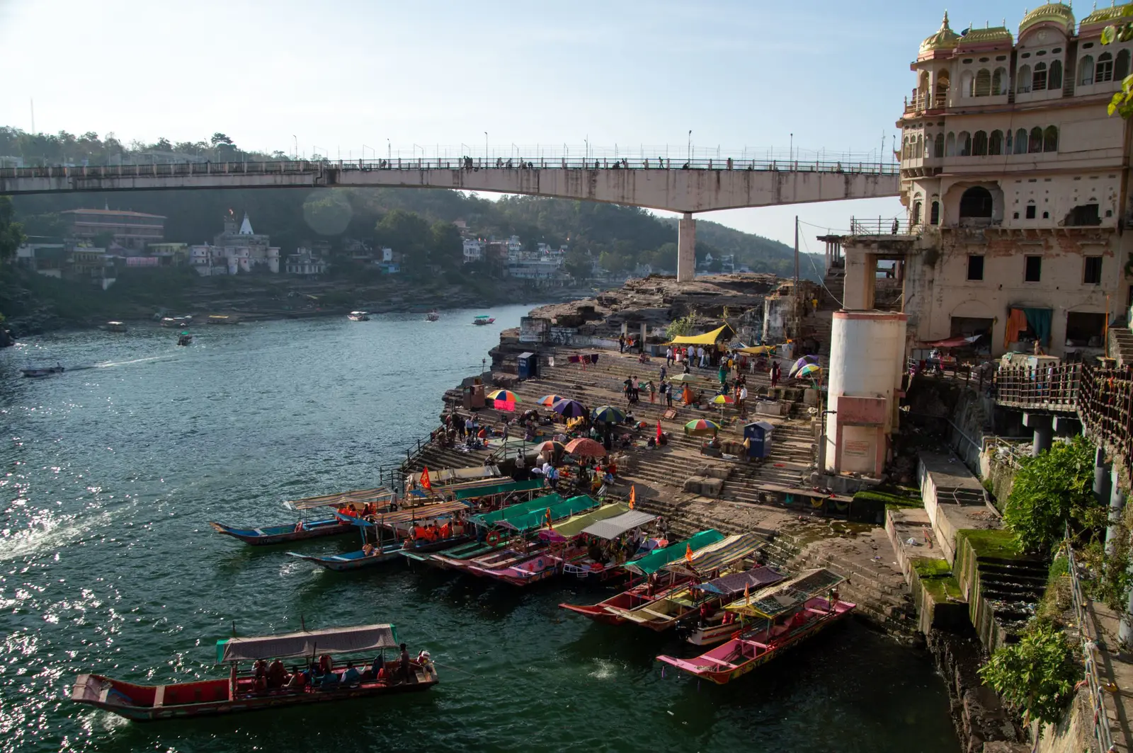 Colourful wooden boats moored at a ghat on the Narmada at Omkareshwar, with a bridge and old riverside buildings behind