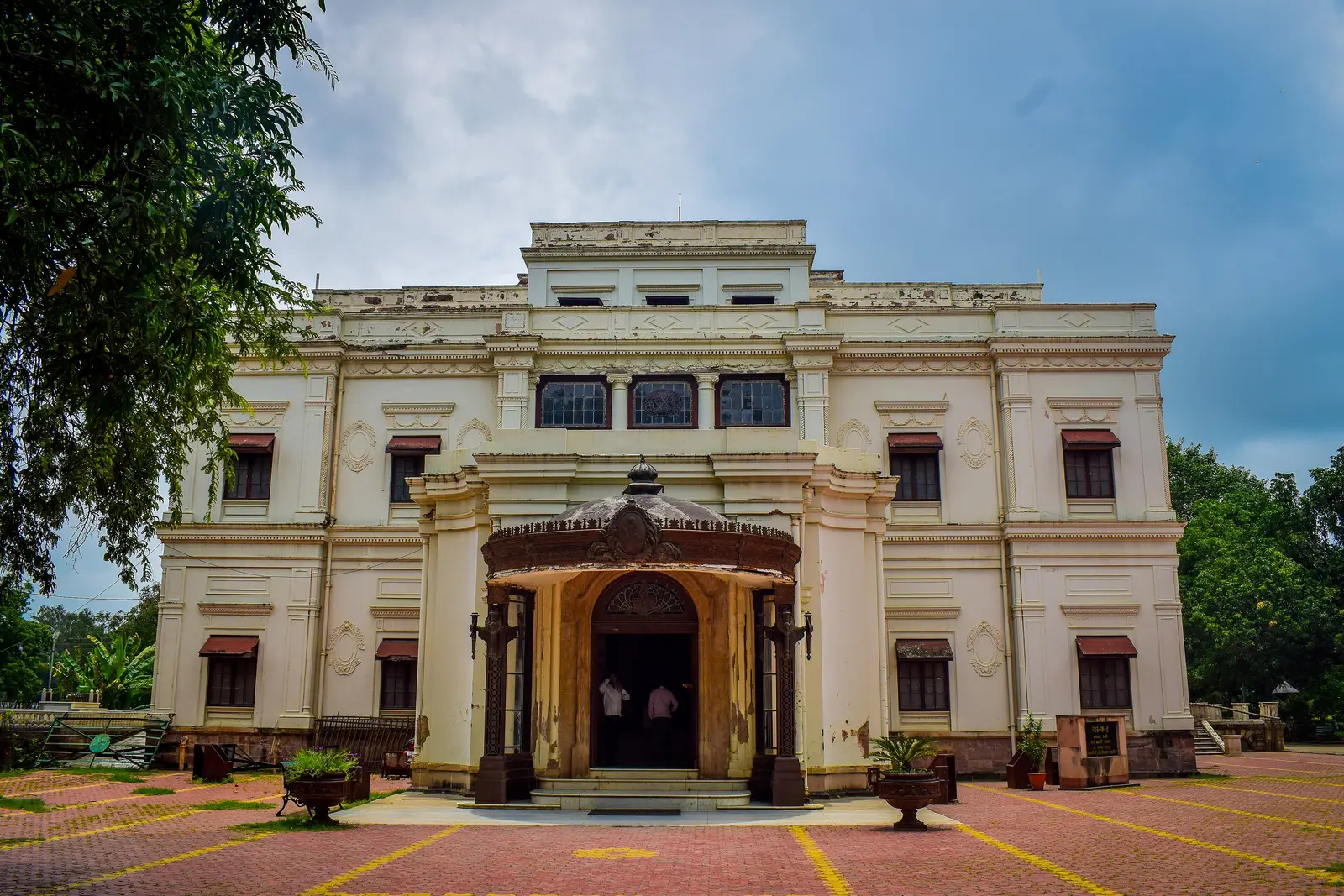 Cream-coloured facade and entrance porch of Lal Bagh Palace in Indore under a cloudy sky, with a paved forecourt
