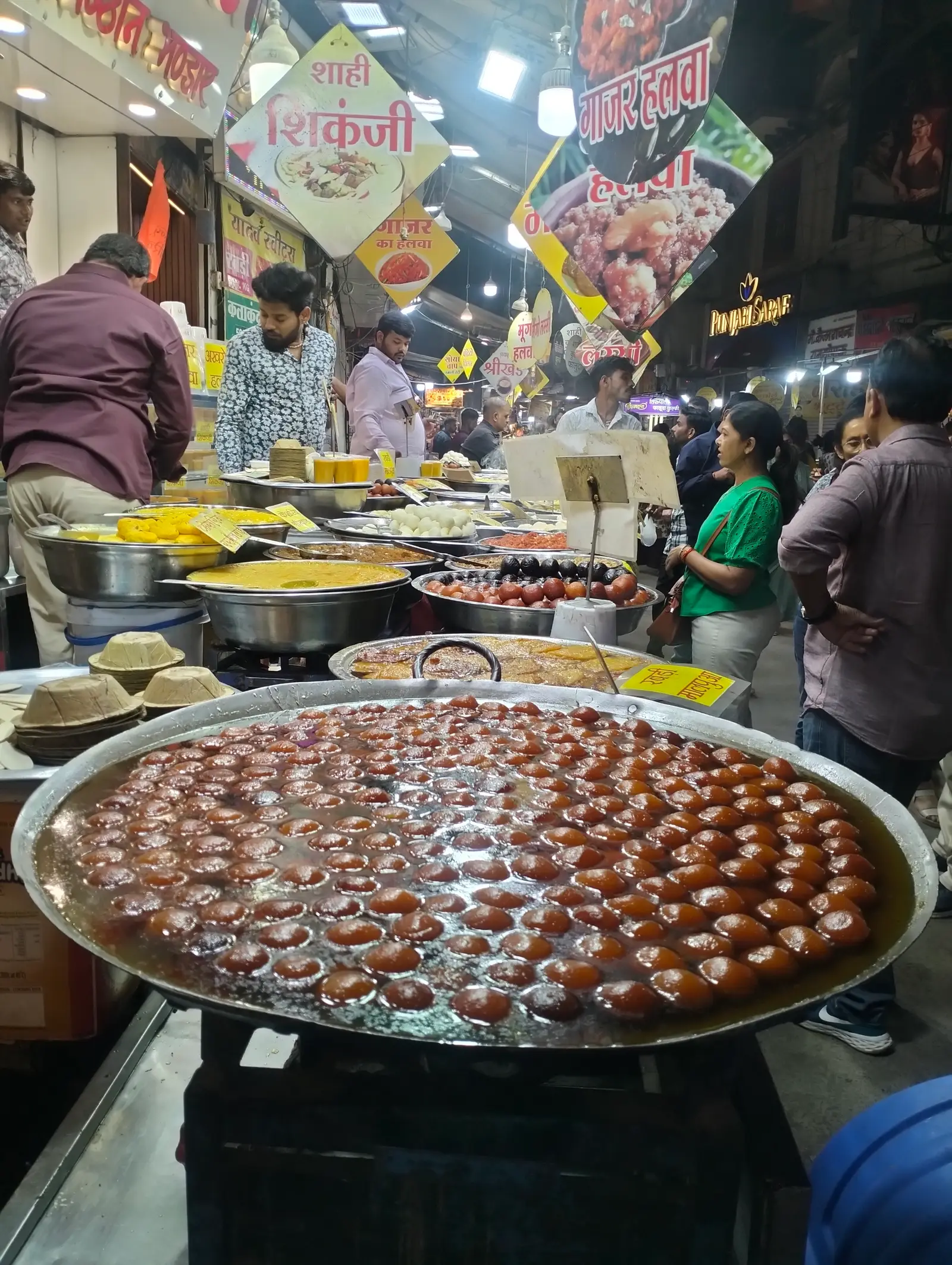 A large kadhai of gulab jamun at a Sarafa Bazaar stall in Indore at night, with vendors, customers and a shikanji signboard behind