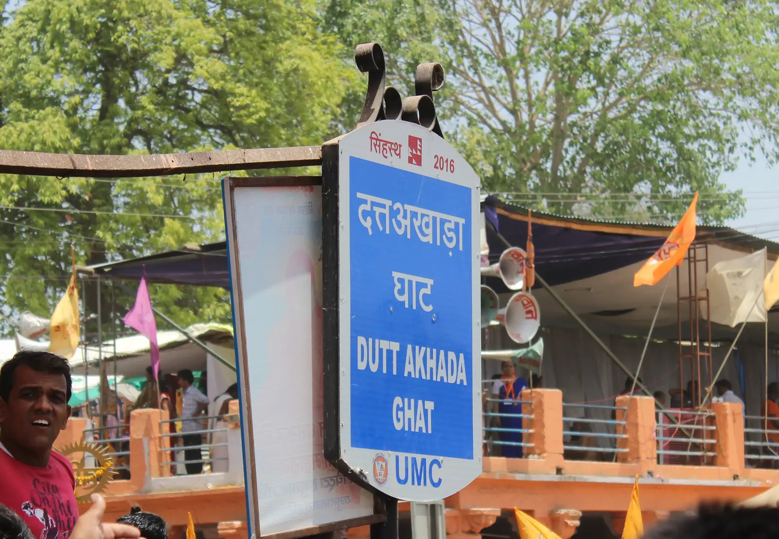 हिंदी और अंग्रेजी में दत्त अखाड़ा घाट लिखा नीला साइनबोर्ड, पीछे भगवा झंडे और नदी किनारे का मंडप, उज्जैन, 2016