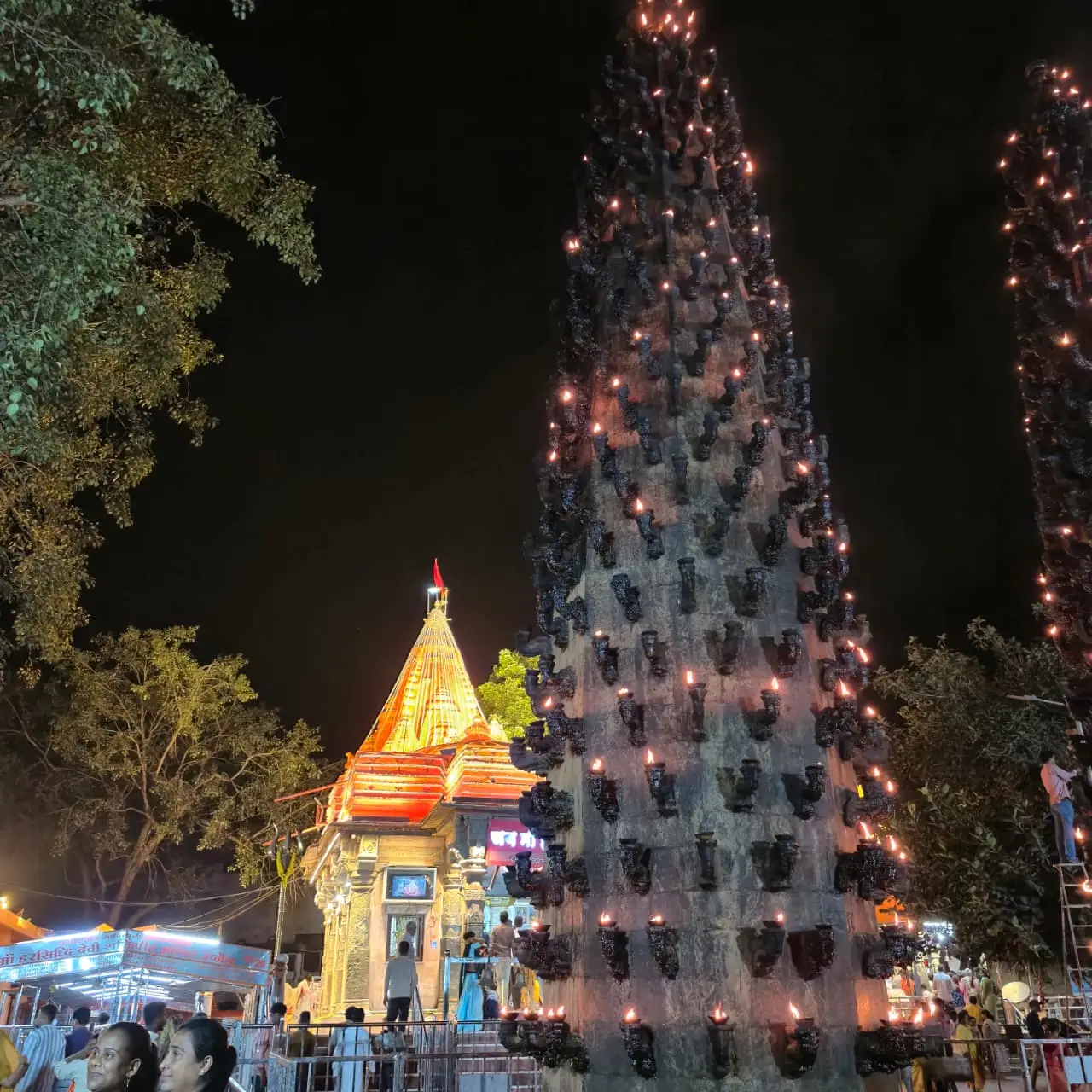 A tall deepstambh lamp pillar covered in lit oil lamps beside the lit shikhara of Harsiddhi Mata temple, Ujjain, at night