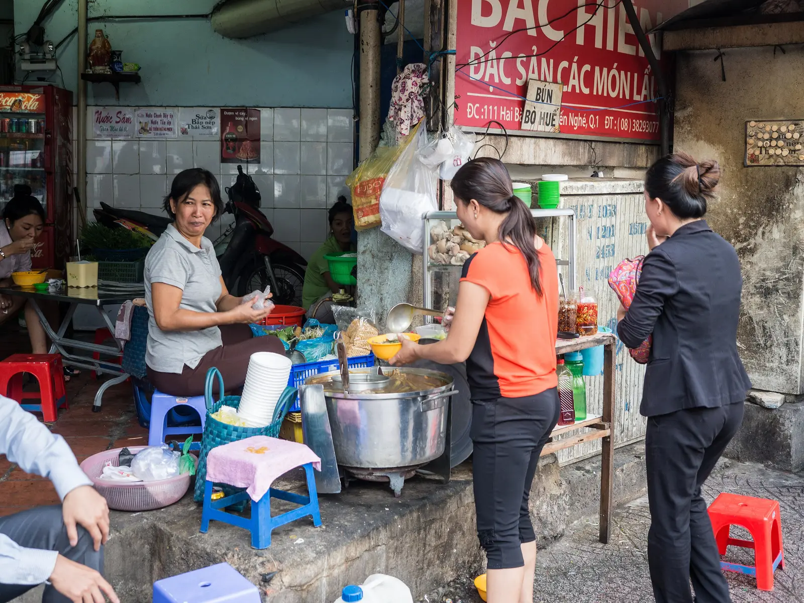 Pavement noodle stall in Vietnam selling bún bò Huế, with a steaming pot, plastic stools and customers ordering